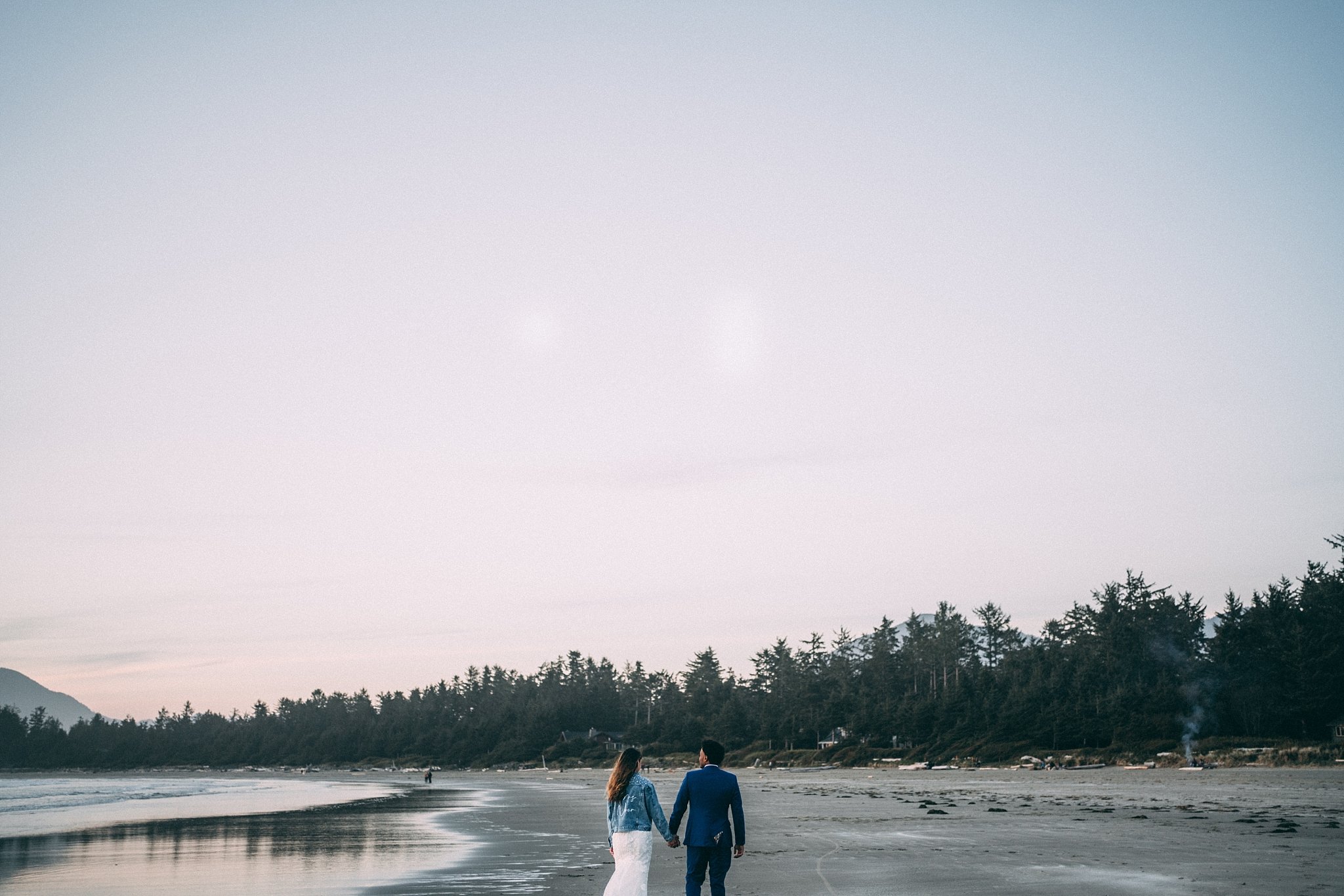 bride in jean jacket groom in blue suit walking on beach