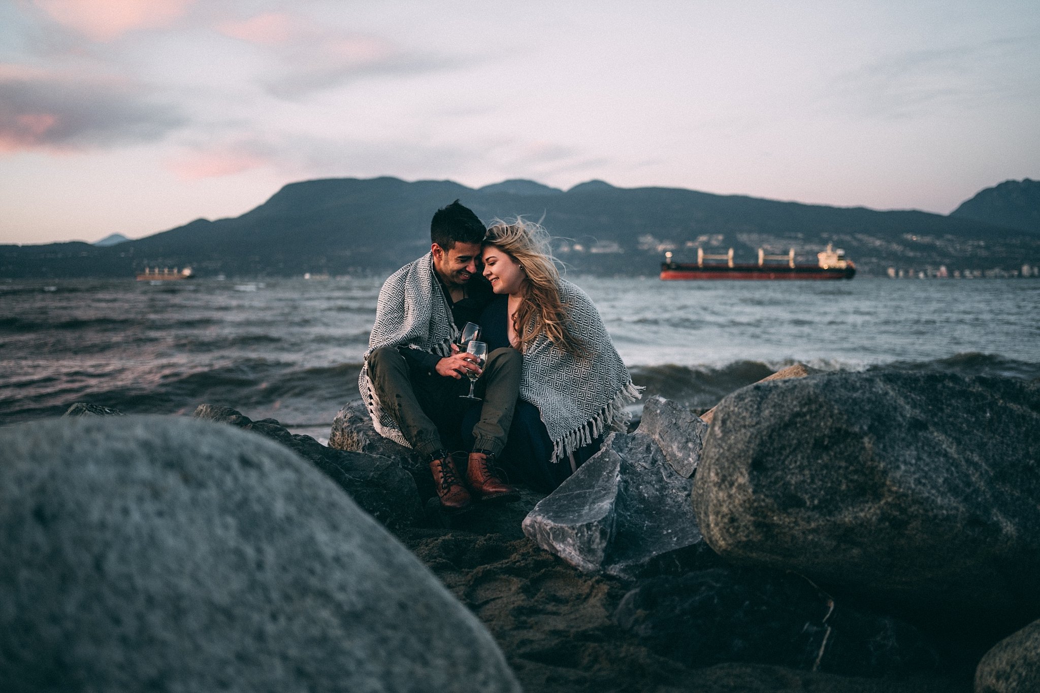 couple cuddling on rocks on vancouver beach with wine glasses